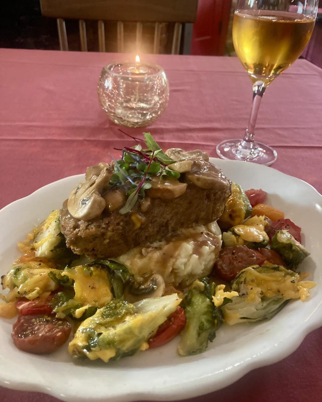 A plate of meatloaf topped with mushrooms on a bed of mashed potatoes and mixed vegetables, with a glass of white wine and a candle in the background.