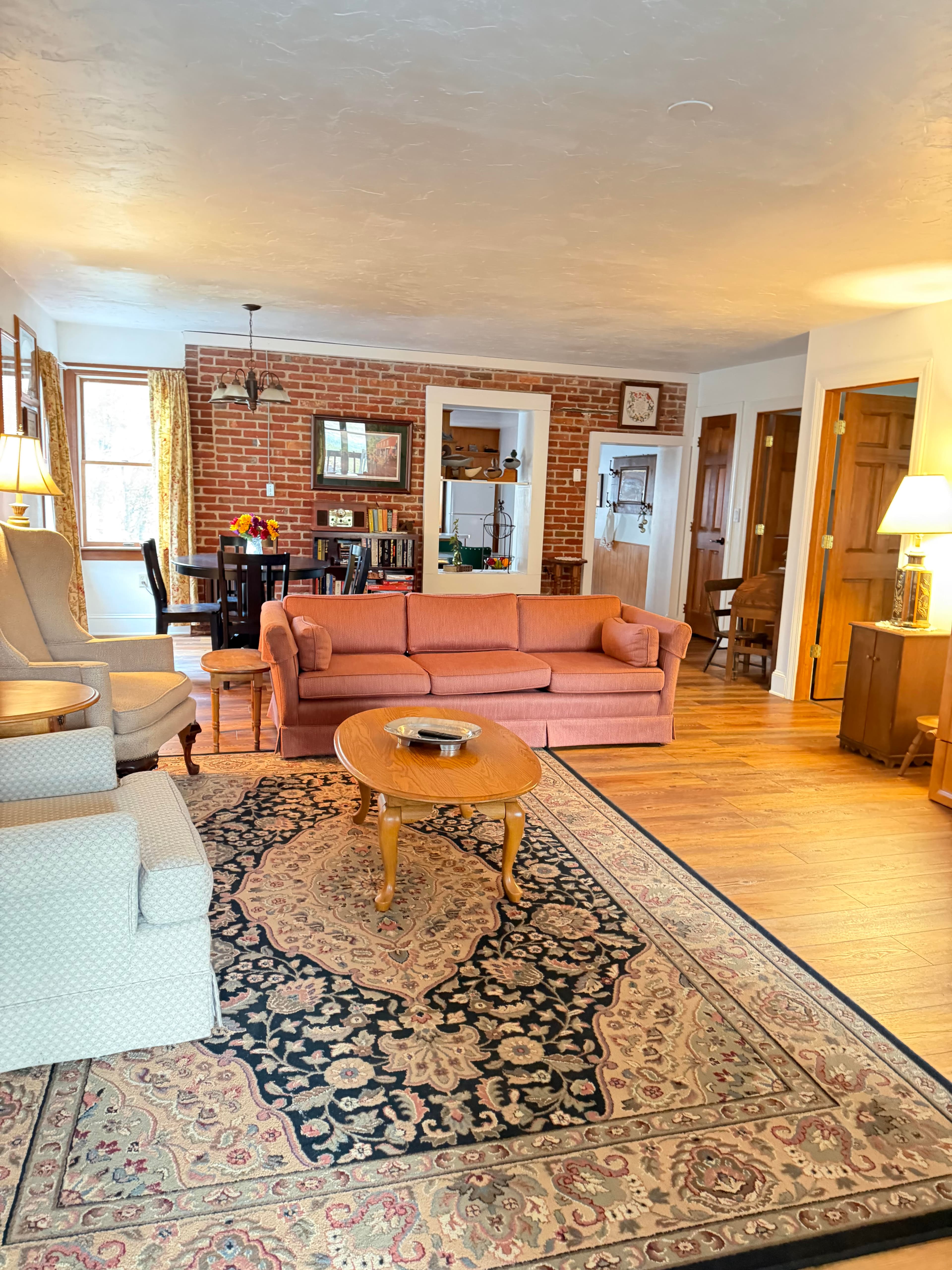 A cozy living room featuring a pink sofa, armchairs, a round coffee table, and exposed brick walls.