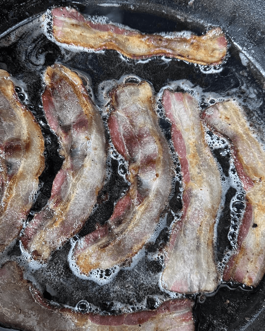 Sizzling strips of bacon frying in a cast iron skillet.
