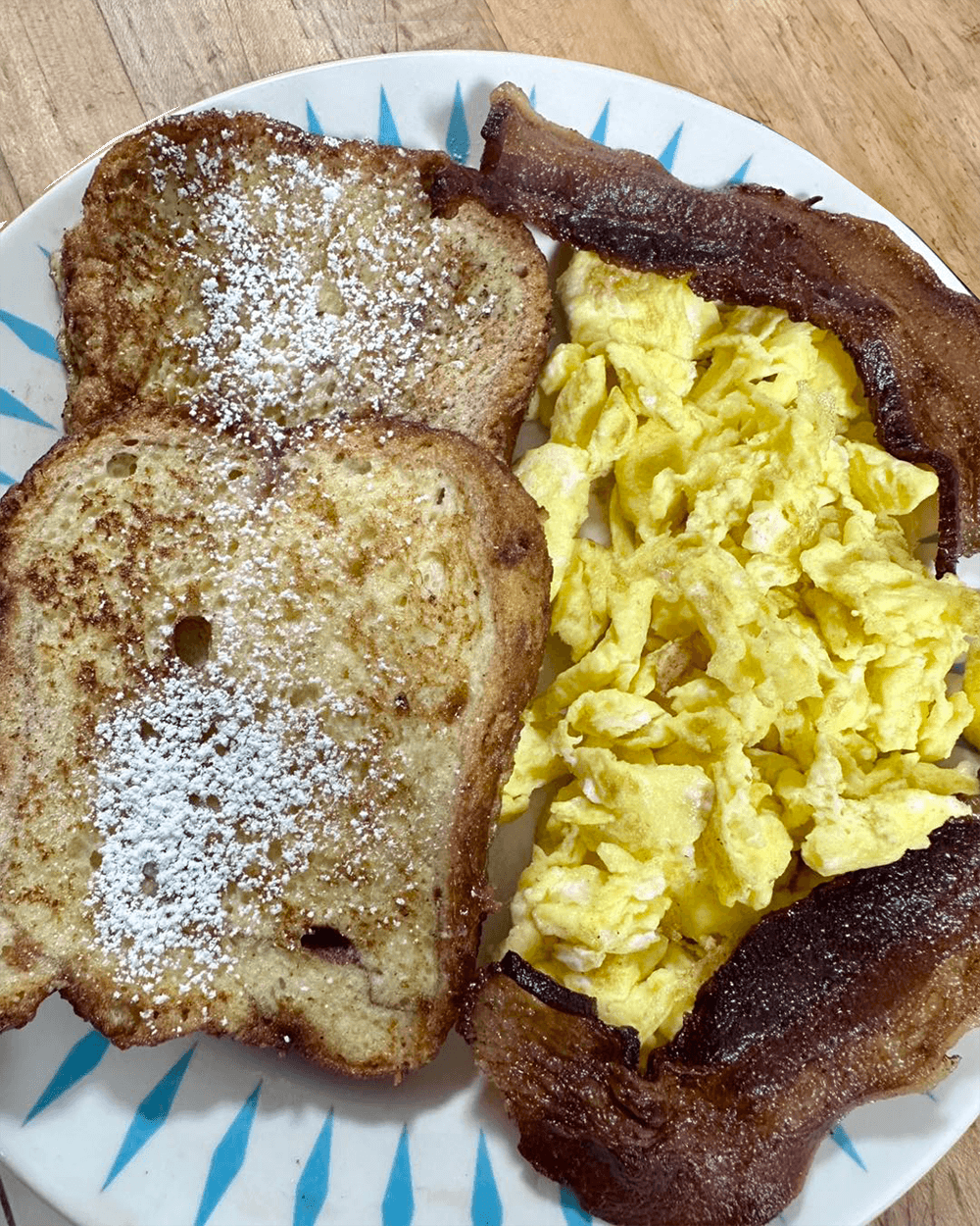 A plate with two slices of French toast dusted with powdered sugar, scrambled eggs, and a slice of bacon.