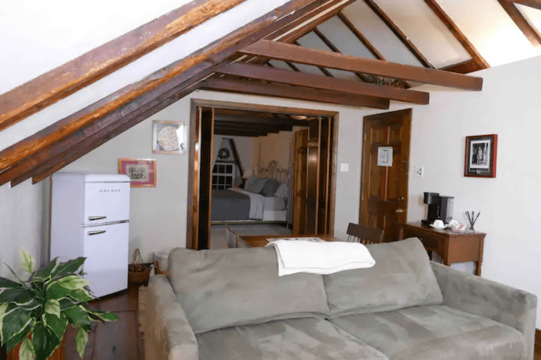 Cozy attic living space featuring a sofa, small fridge, and a glimpse of a bedroom through an open doorway.