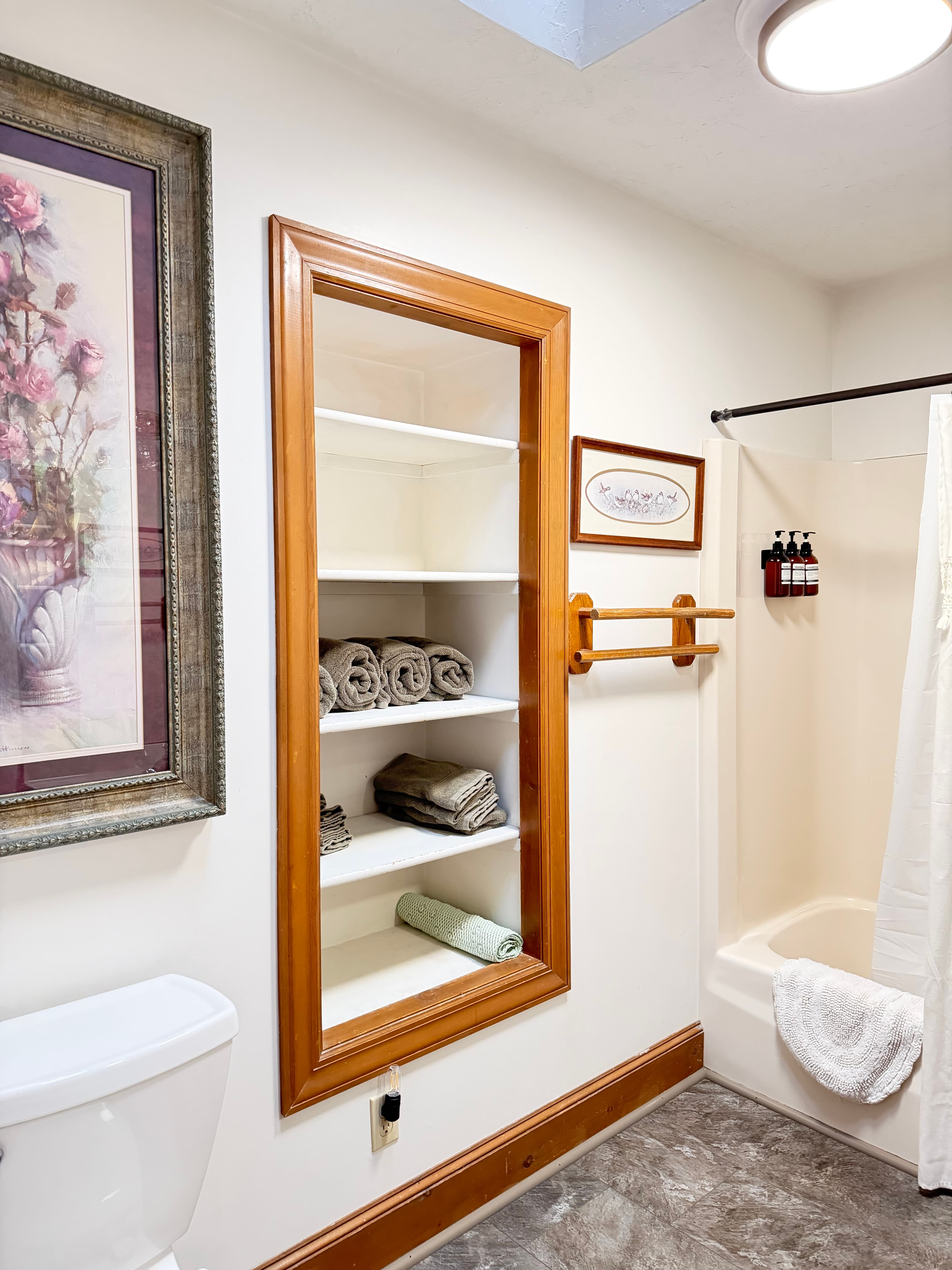 A well-lit bathroom featuring a shower, a mirror with shelves, and neatly arranged towels.