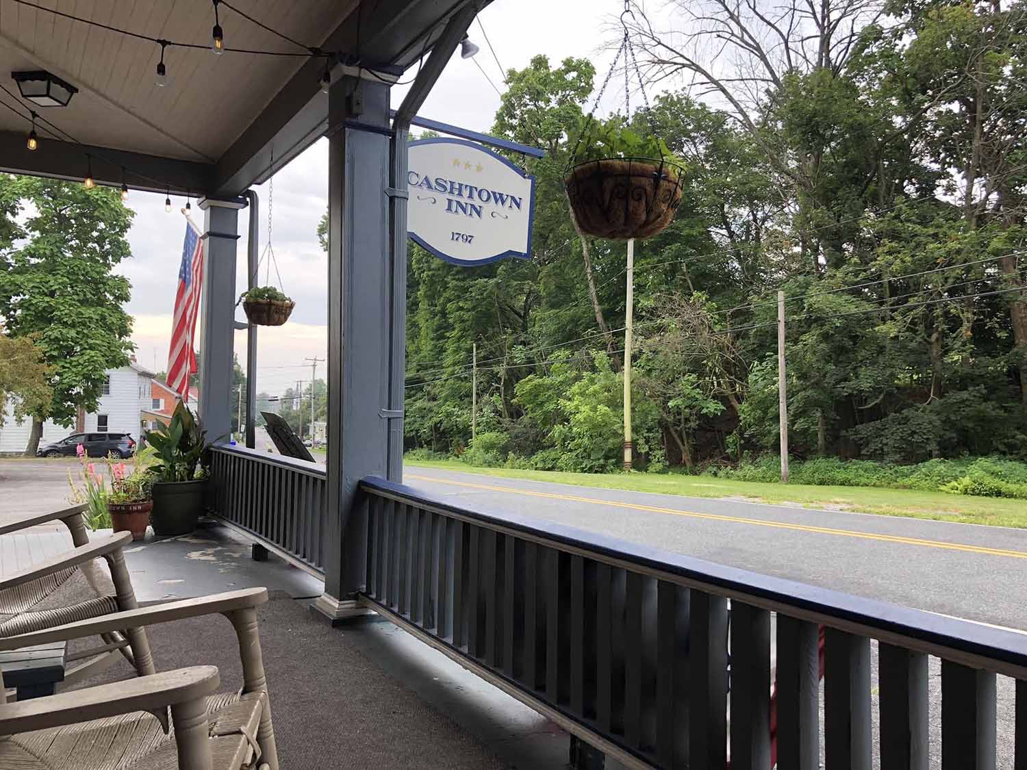 View of a porch with rocking chairs, featuring a hanging sign for Cashtown Inn and an American flag, overlooking a tree-lined road.