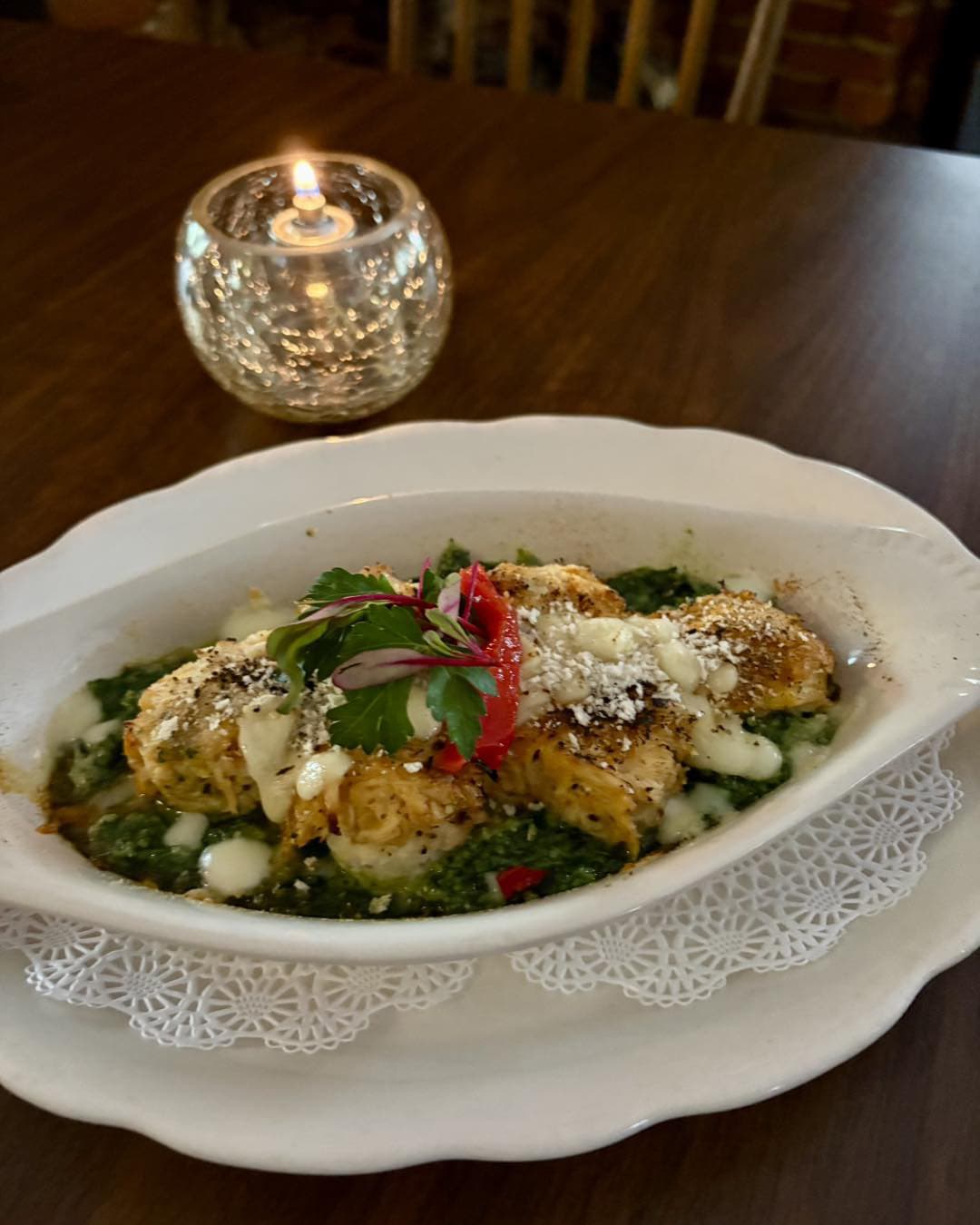 A dish of savory baked fish served on a bed of green sauce, garnished with herbs and a candle in the background.
