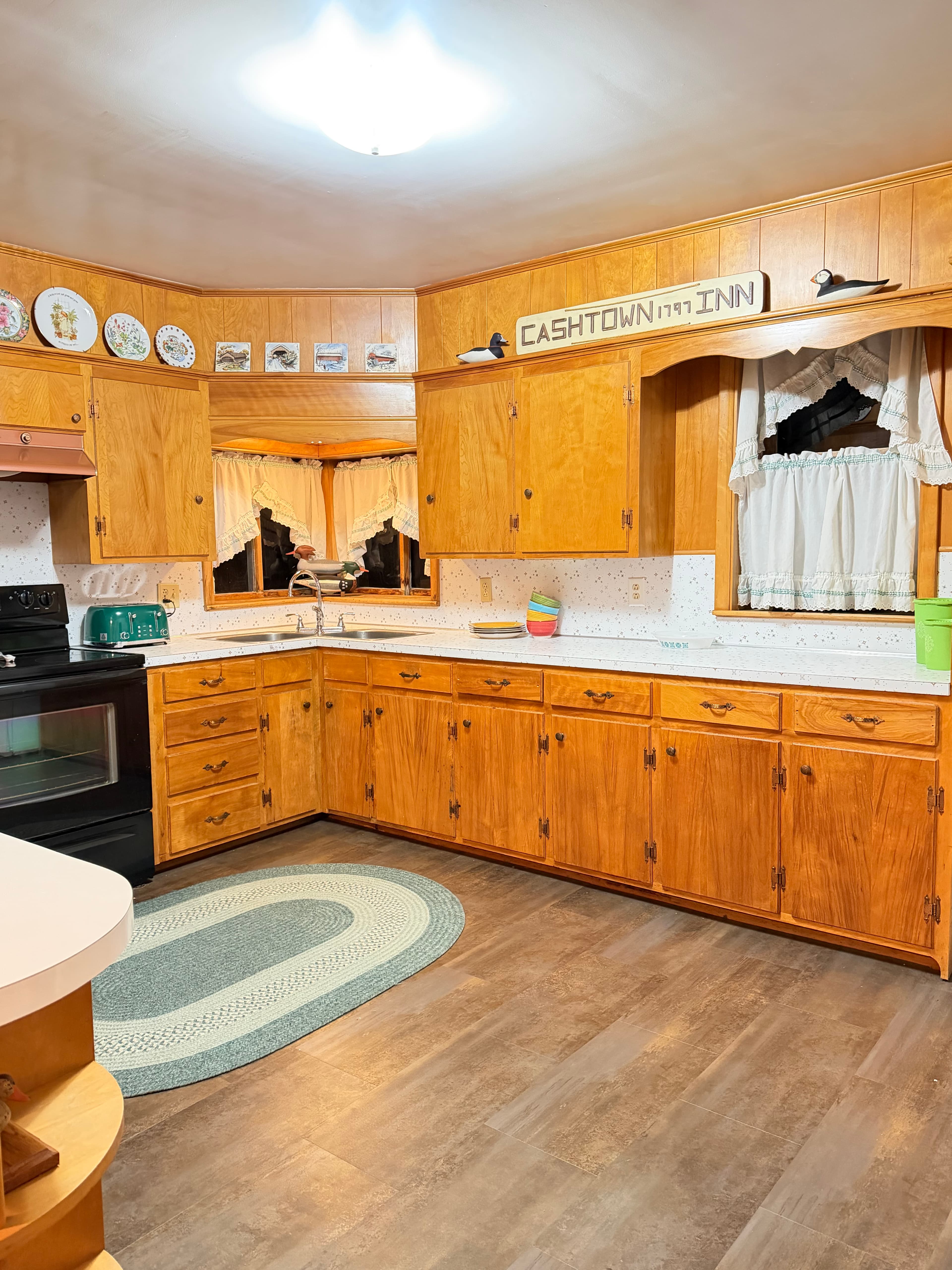A warm, wood-paneled kitchen featuring a black stove, a window with white curtains, and decorative plates on the wall.