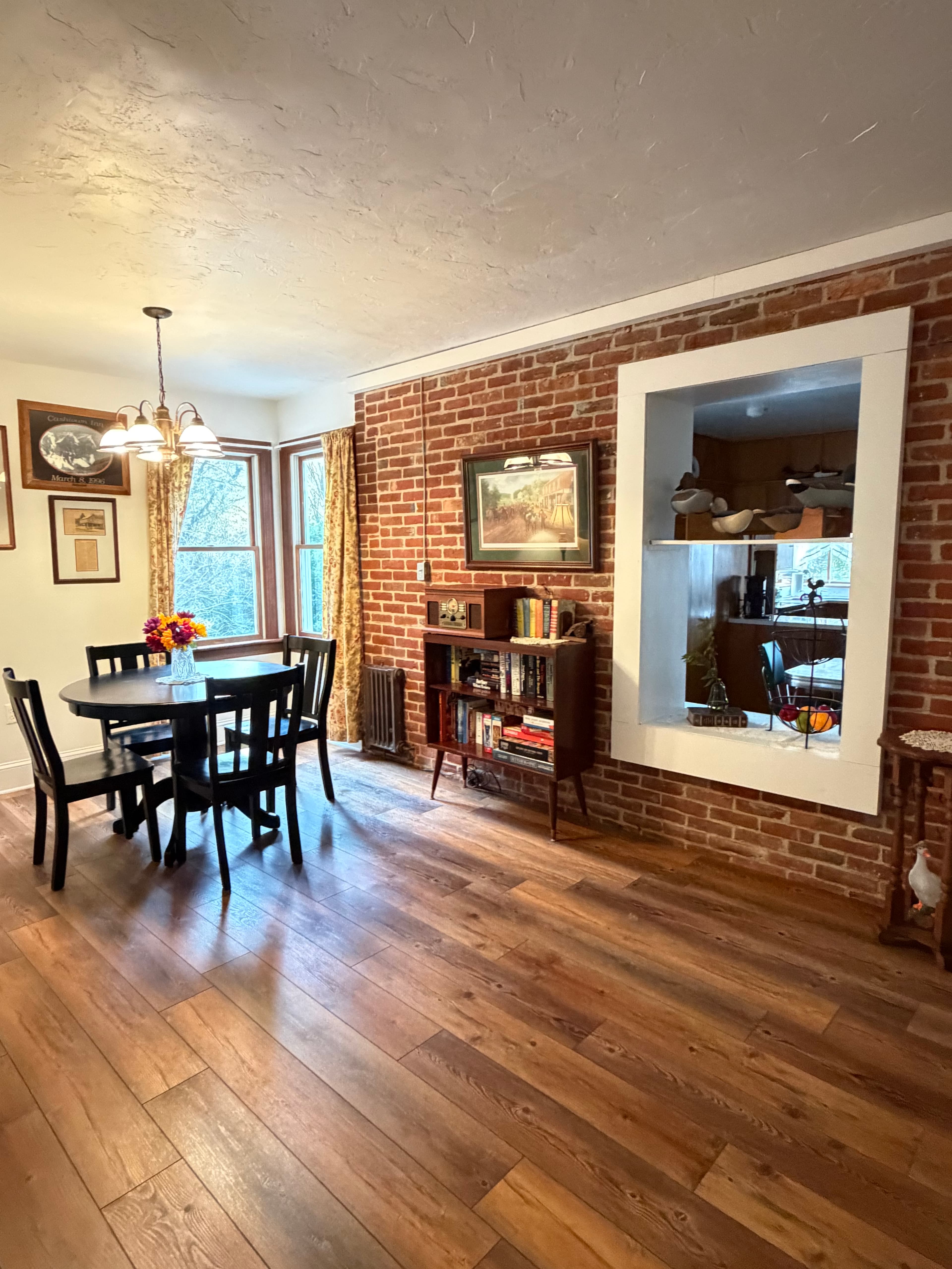 A cozy dining area features a wooden table, brick walls, and large windows with curtains.