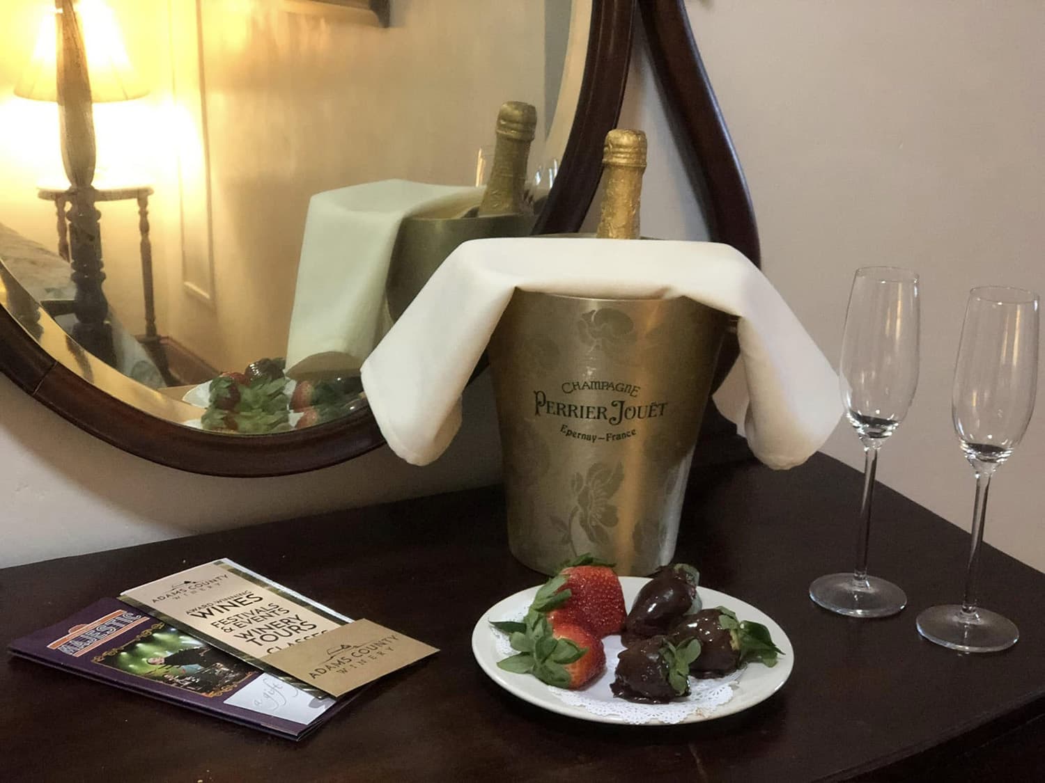 A bottle of Perrier-Jouët champagne in an ice bucket, accompanied by chocolate-covered strawberries and two champagne flutes, sits on a wooden table beside a mirrored wall.