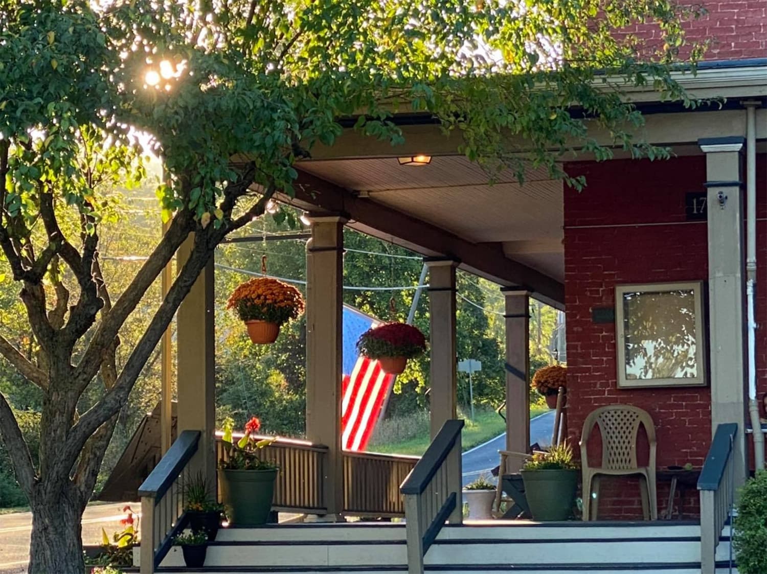 A cozy porch with potted plants and an American flag hanging, framed by trees and soft sunlight.