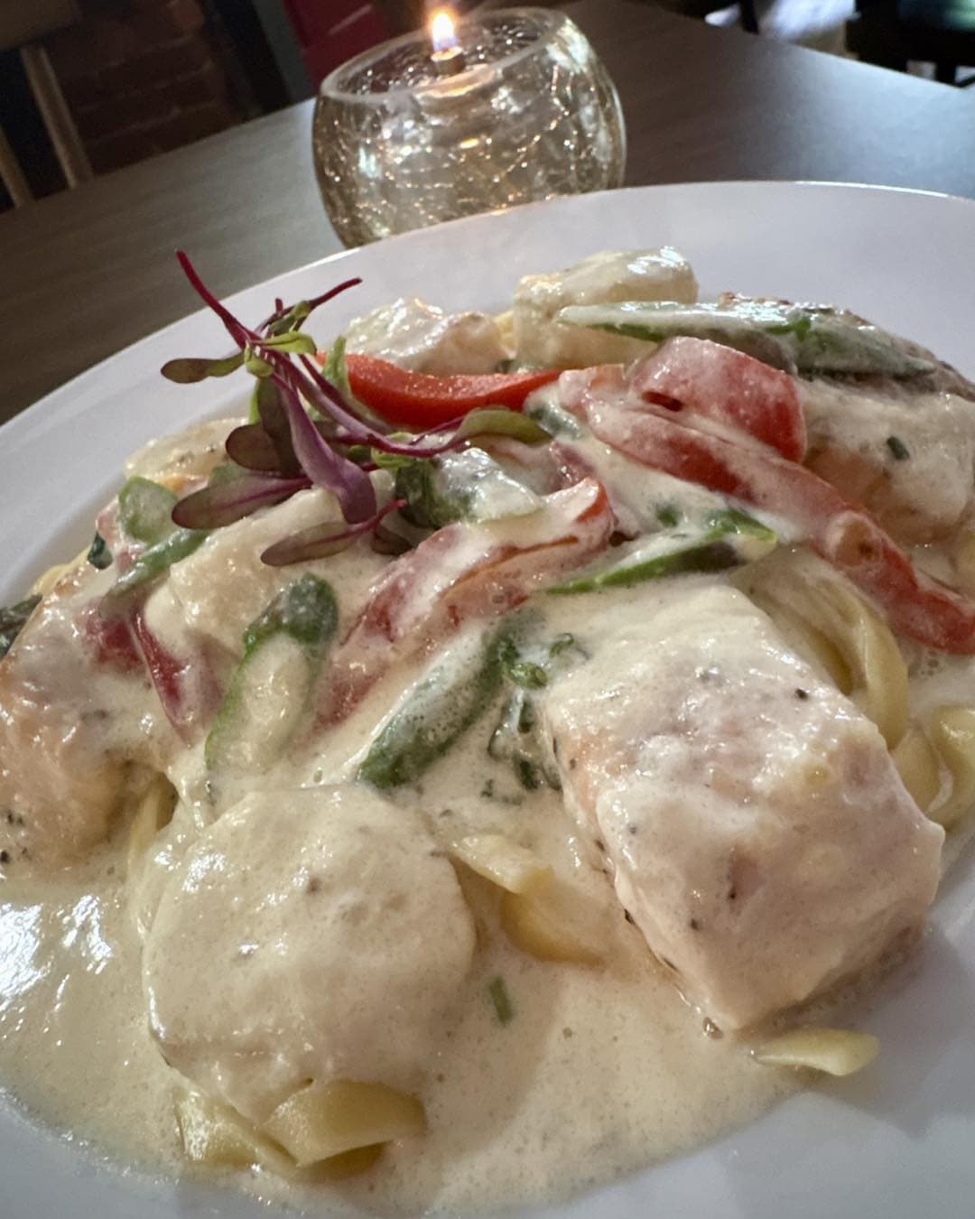 Creamy pasta with chicken, bell peppers, and greens, garnished with microgreens, next to a lit candle.