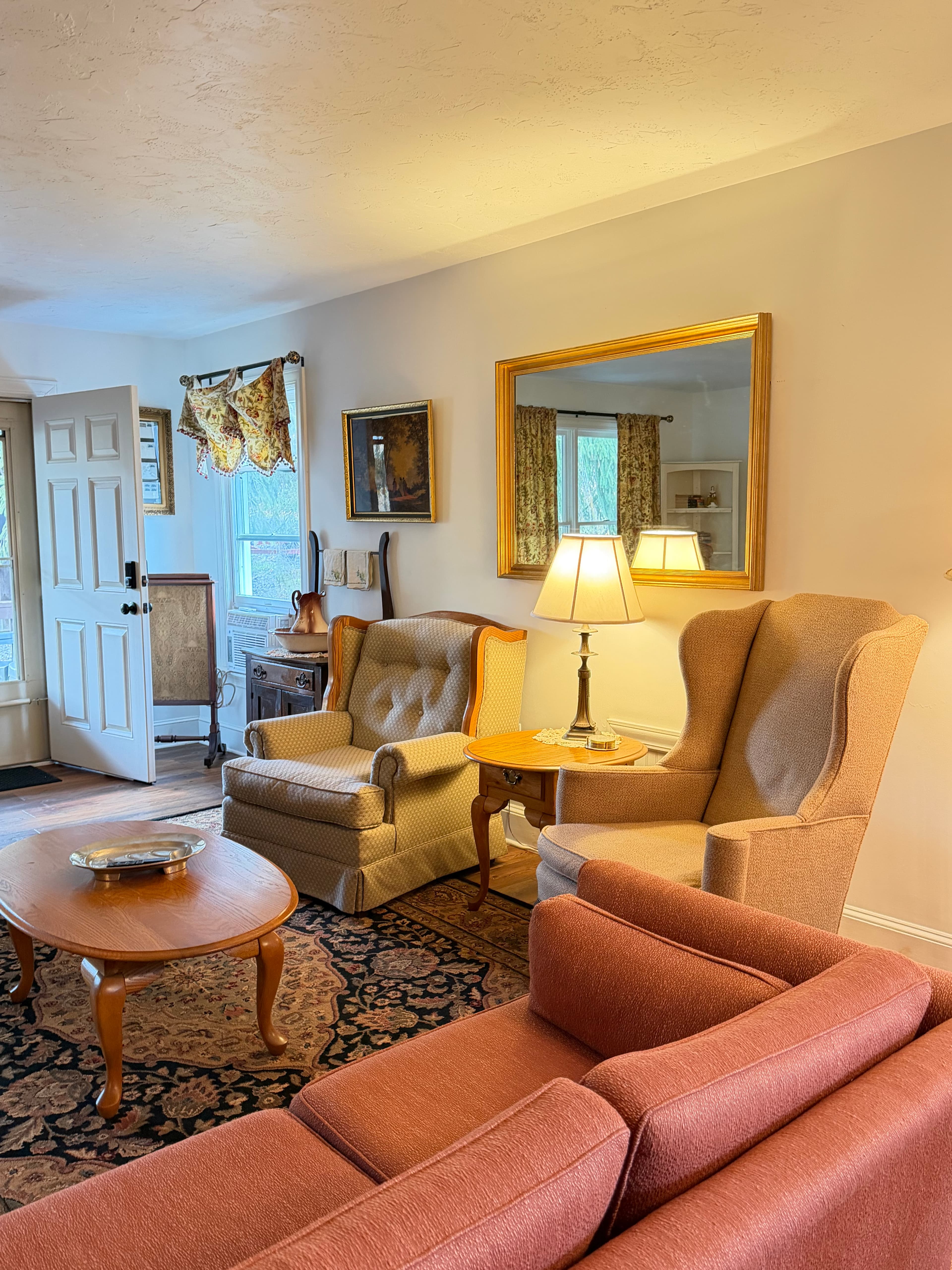 A cozy living room featuring two armchairs, a coffee table, and a lamp, with natural light coming from an open door.