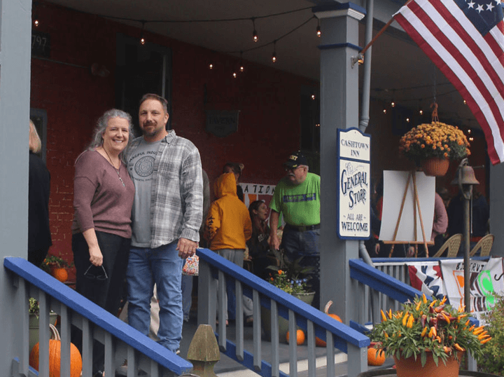 A couple poses on the porch of a general store decorated with autumn pumpkins and flowers.