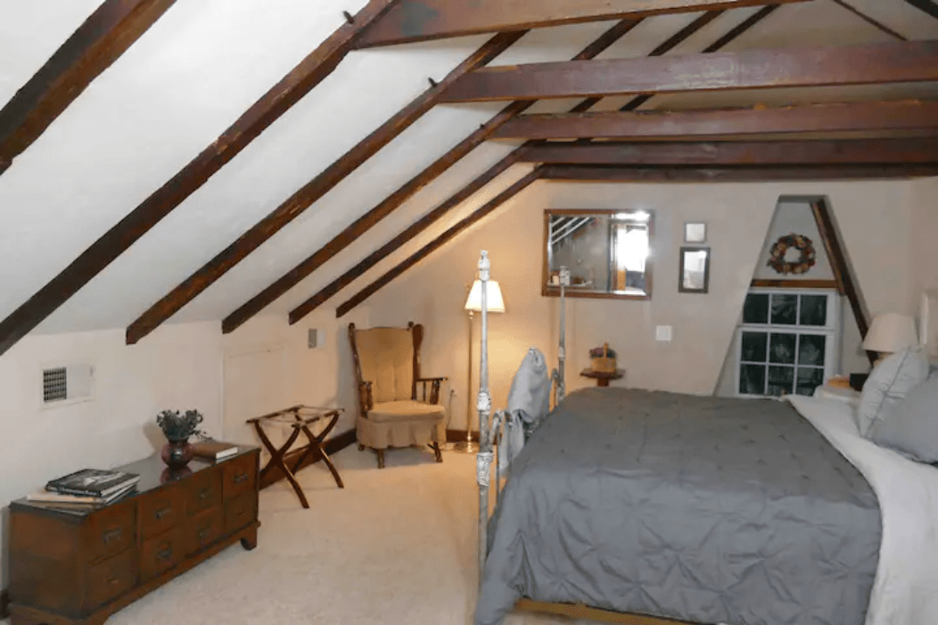 Cozy attic bedroom featuring a sloped ceiling, wooden beams, a bed, and a reading nook with a chair and lamp.