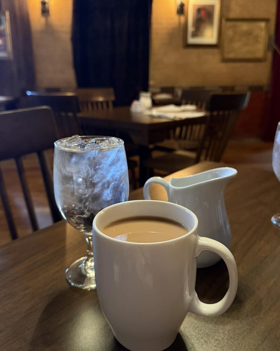 A white coffee mug filled with coffee sits beside a glass of ice water and a small cream pitcher on a wooden table in a cozy restaurant.