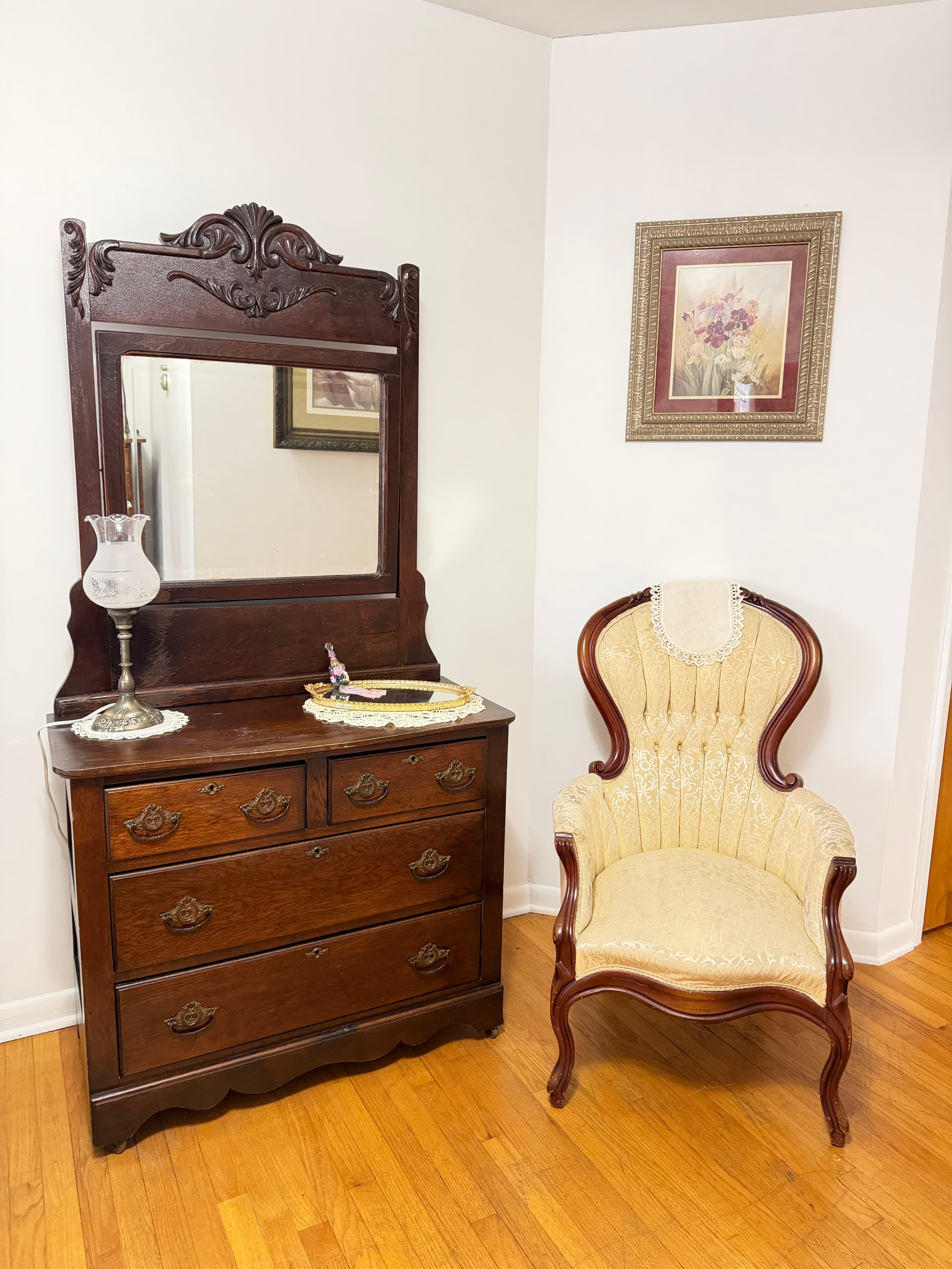 A wooden dresser with a mirror and a vintage upholstered chair beside it, set against a white wall.