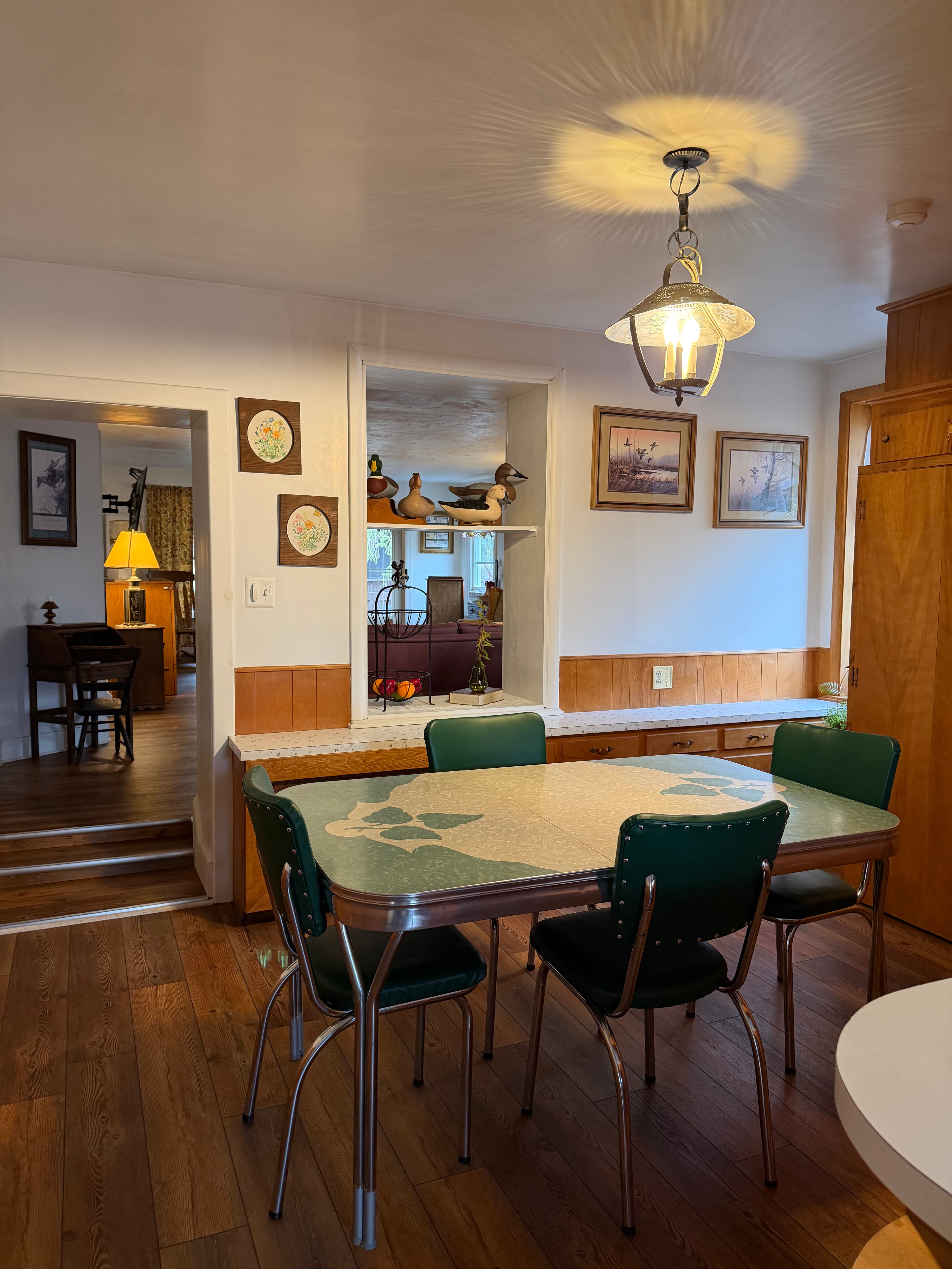 A cozy kitchen featuring a retro dining table with green chairs, warm wooden accents, and framed artwork on the walls.