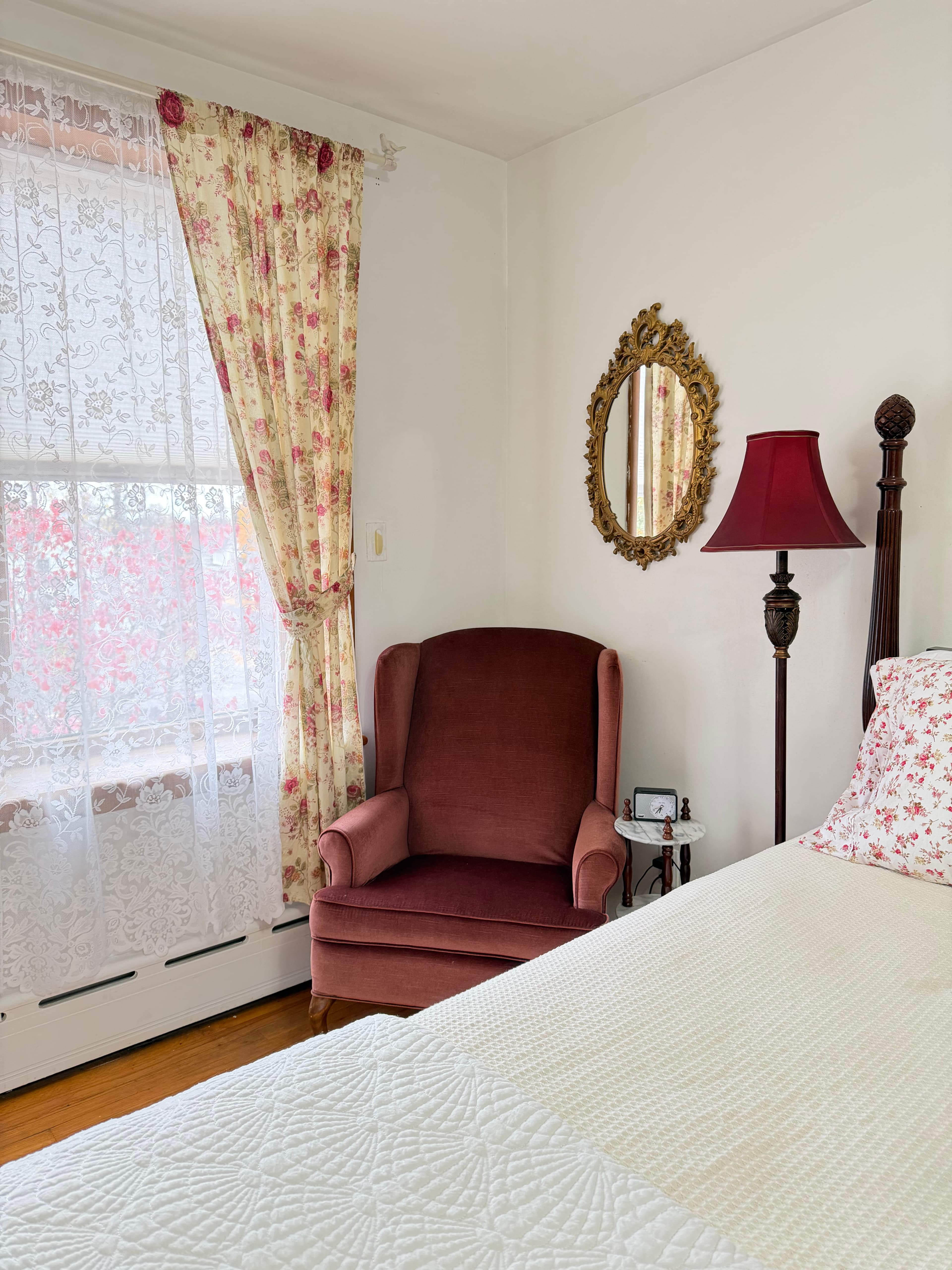 A cozy bedroom featuring a red armchair, floral curtains, a round mirror, and a lamp beside a bed.