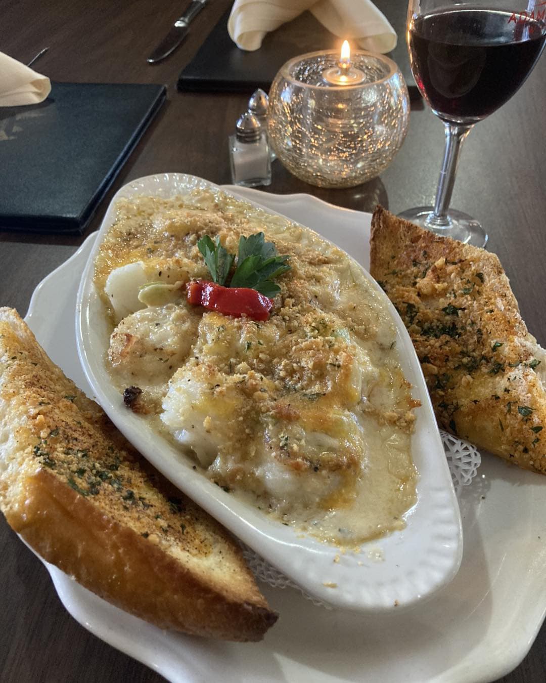 A creamy seafood dish topped with breadcrumbs, served with garlic bread and accompanied by a glass of red wine.