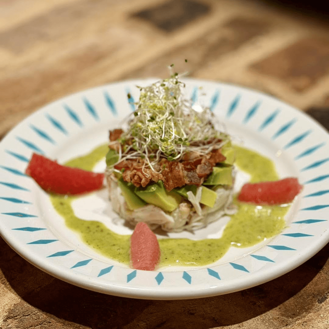 A beautifully plated dish featuring avocado, bacon, and sprouts on a bed of crab, garnished with grapefruit and a green sauce.